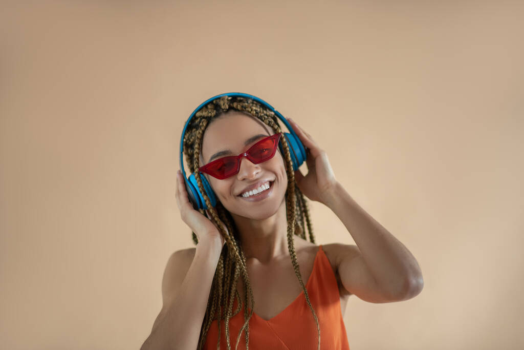 Улыбающаяся молодая афроамериканка в синих наушниках и красных очках, слушающая музыку
 - Фото, изображение