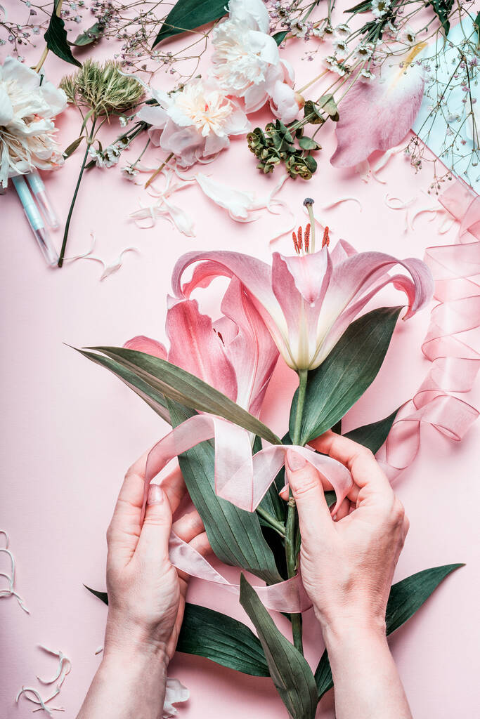 Weibliche Hände machen schöne rosa Lilienblüten Strauß auf pastellfarbenem Tisch mit Florist Dekoration Ausrüstung, Draufsicht. Kreativer Floristenarbeitsplatz und Blumenarrangements. Festliches Ferienkonzept. - Foto, Bild