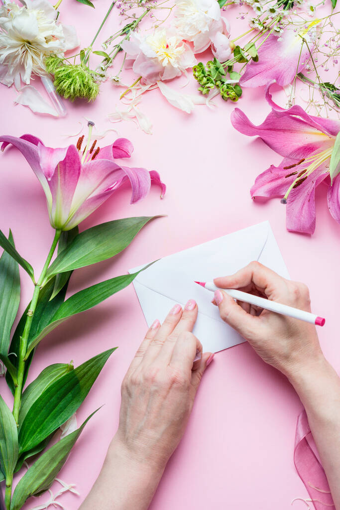 Female hands write with pencil greeting card on Blank envelop on pink table background with lily flowers and florist decoration equipment, top view. Creative  Invitation and holiday  concept - Photo, Image