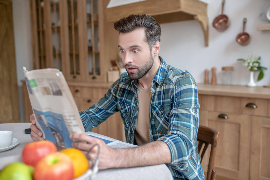 Мужчина в клетчатой рубашке сидит за столом, читает газету и выглядит удивлённым
 - Фото, изображение