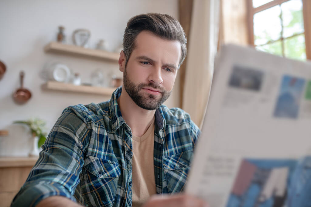 Man in een geruit shirt zit aan tafel, leest kranten en ziet er serieus uit. - Foto, afbeelding