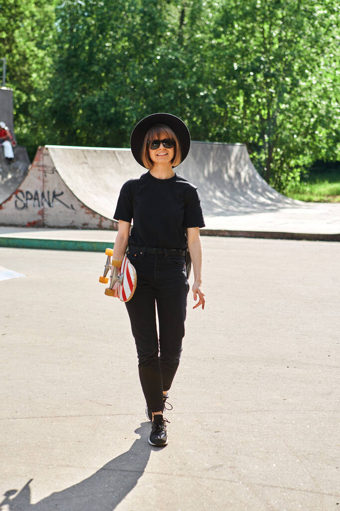 Śliczna uśmiechnięta kobieta niesie longboard spacerując w skateparku w okularach przeciwsłonecznych, kapeluszu i czarnych ubraniach - Zdjęcie, obraz