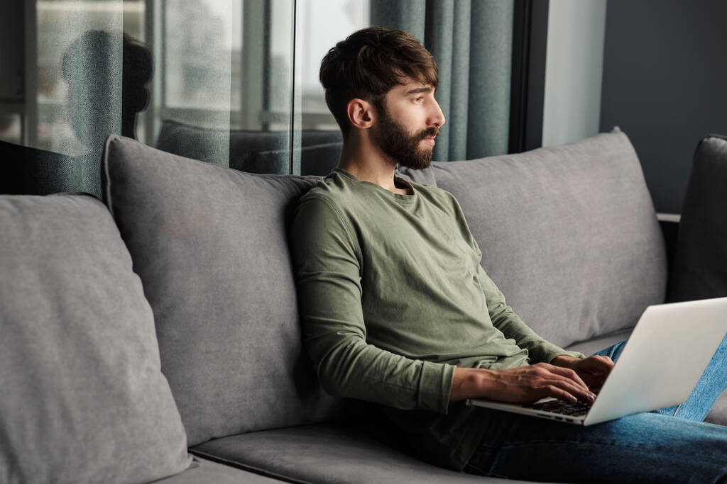 Изображение ориентированного молодого человека в повседневной одежде с помощью ноутбука, сидящего на диване в гостиной
 - Фото, изображение