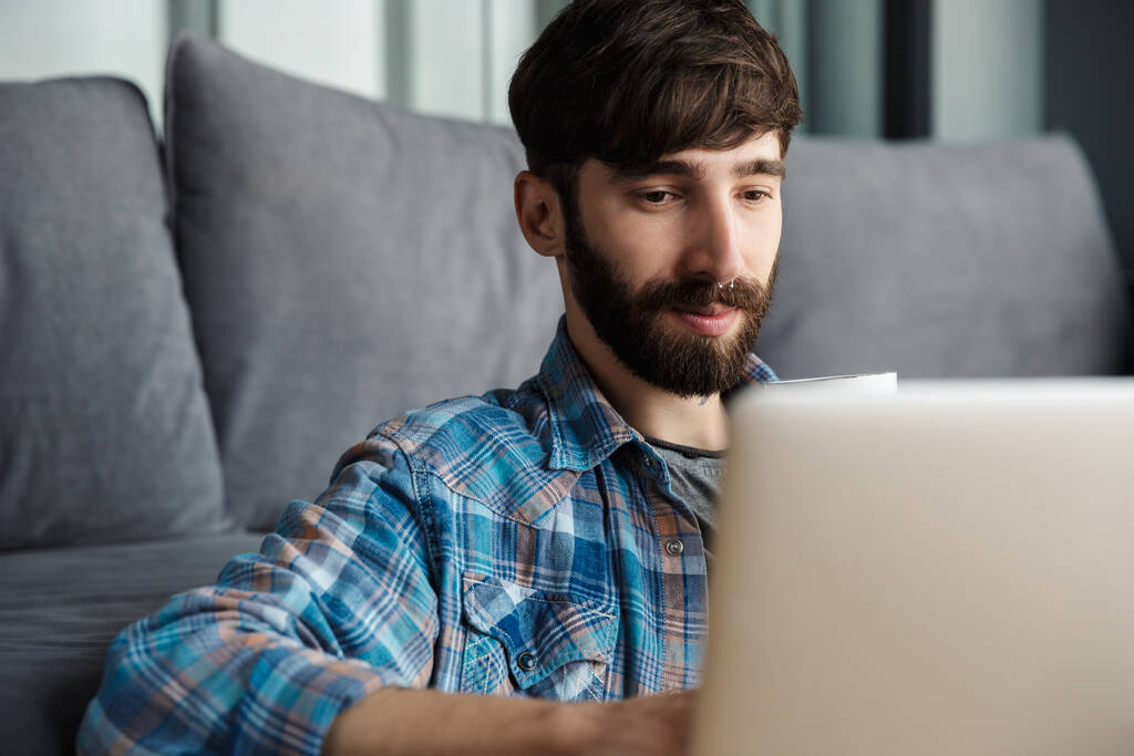 Image d'un homme barbu pensant travaillant avec un ordinateur portable assis sur le sol dans le salon - Photo, image
