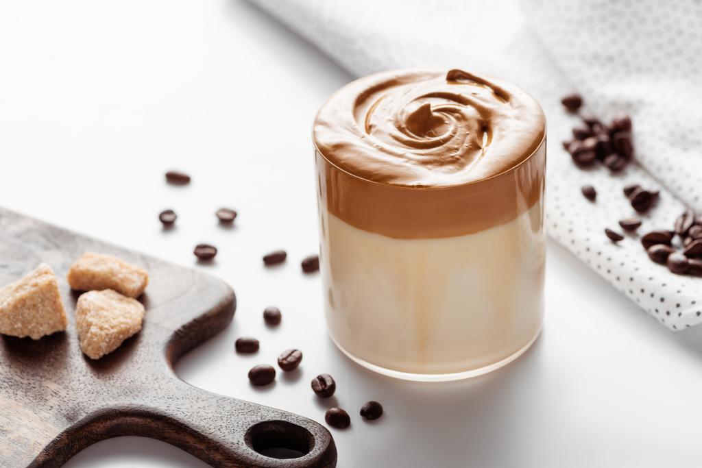 selektiver Fokus von köstlichem Dalgona-Kaffee im Glas in der Nähe von Kaffeebohnen, braunem Zucker auf Schneidebrett und Serviette auf weißem Hintergrund - Foto, Bild