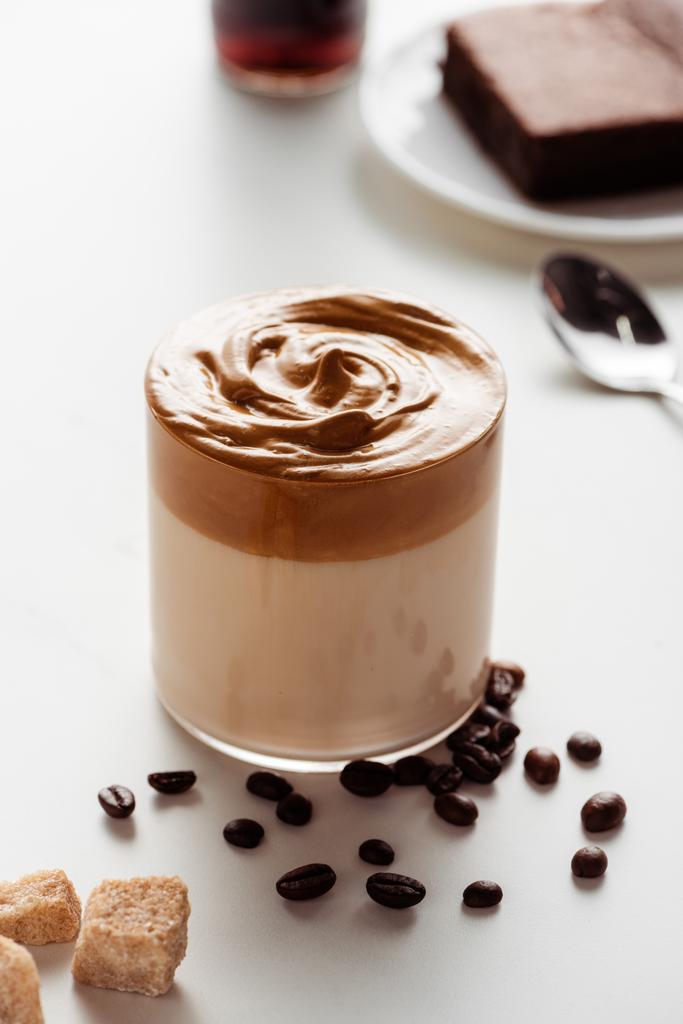 herkullisen Dalgona-kahvin valikoiva keskittyminen lasiin lähellä kahvipapuja ja ruskeaa sokeria valkoisella taustalla - Valokuva, kuva