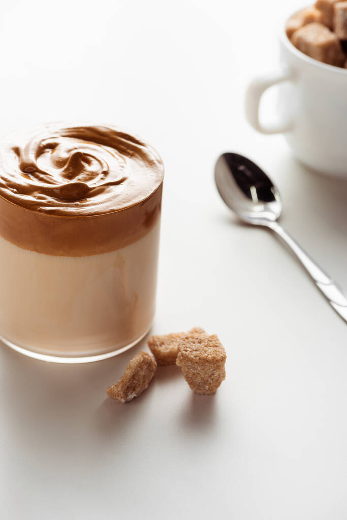 селективный фокус вкусного кофе Dalgona в стакане возле ложки и коричневый сахар на белом фоне
 - Фото, изображение