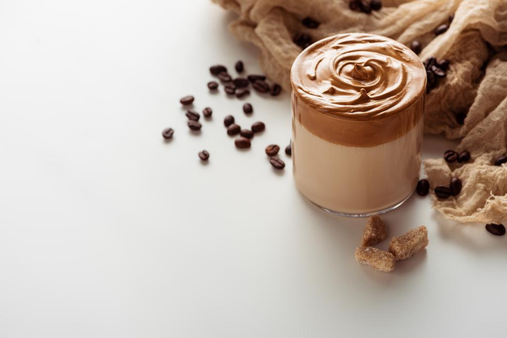 вкусный кофе Dalgona в стекле возле кофейных зерен, ткани и гранулированного коричневого сахара на белом фоне
 - Фото, изображение