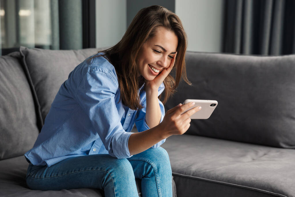 Зображення красивої веселої милої жінки, яка посміхається і використовує мобільний телефон, сидячи на дивані у вітальні
 - Фото, зображення