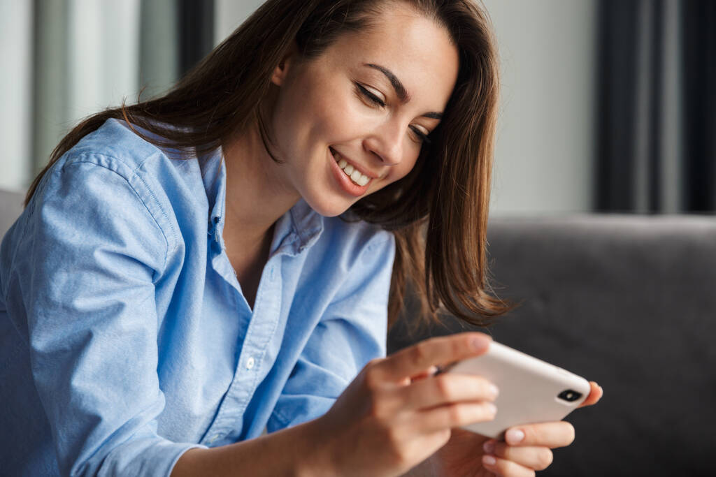 Imagen de una hermosa mujer alegre jugando videojuegos en el teléfono móvil mientras está sentada en la sala de estar
 - Foto, imagen
