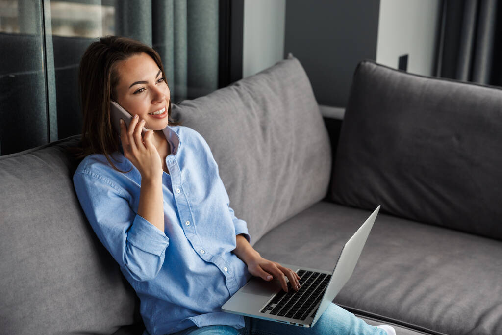Obraz młodej uśmiechniętej kobiety rozmawiającej przez telefon komórkowy i korzystającej z laptopa podczas siedzenia w salonie - Zdjęcie, obraz
