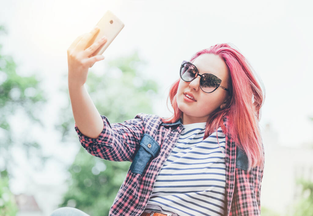 Mooie moderne glimlachende jonge vrouwelijke tiener met buitengewone kapsel kleur in een geruite shirt en zonnebril nemen selfie met behulp van moderne smartphone. Leuke jeugd dagelijks leven en tieners concept - Foto, afbeelding