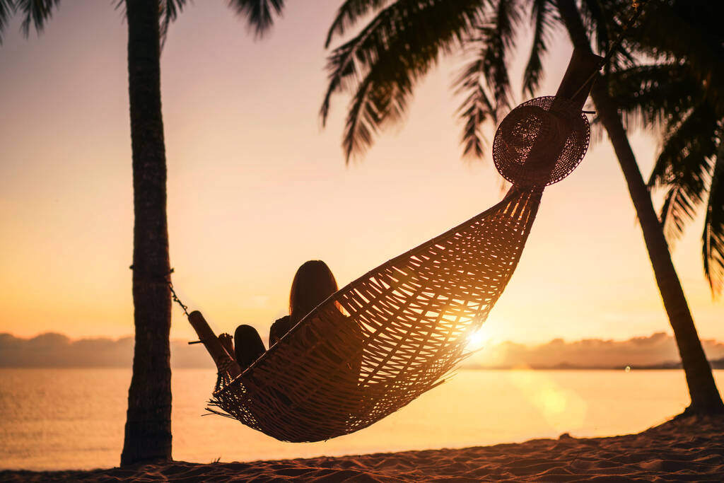Giovane donna rilassante in amaca incernierata tra le palme sulla spiaggia di sabbia all'alba arancione - Foto, immagini