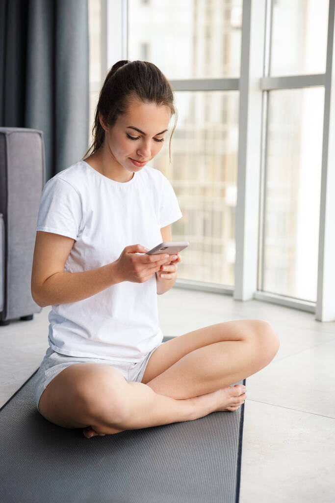 Зображення усміхненої молодої жінки, що використовує мобільний телефон, сидячи на килимку йоги біля вікна в приміщенні
 - Фото, зображення