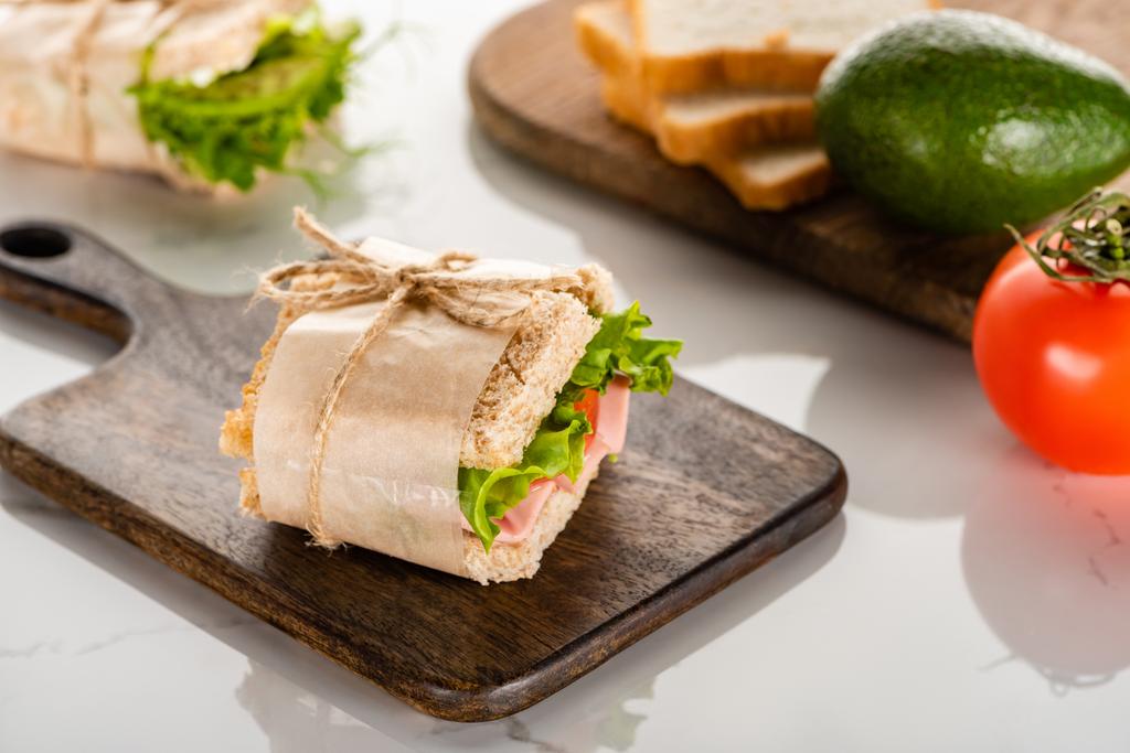Taze yeşil sandviçin seçici odağı ahşap kesim tahtası yanında domates ve beyaz mermer yüzeyde avokado. - Fotoğraf, Görsel