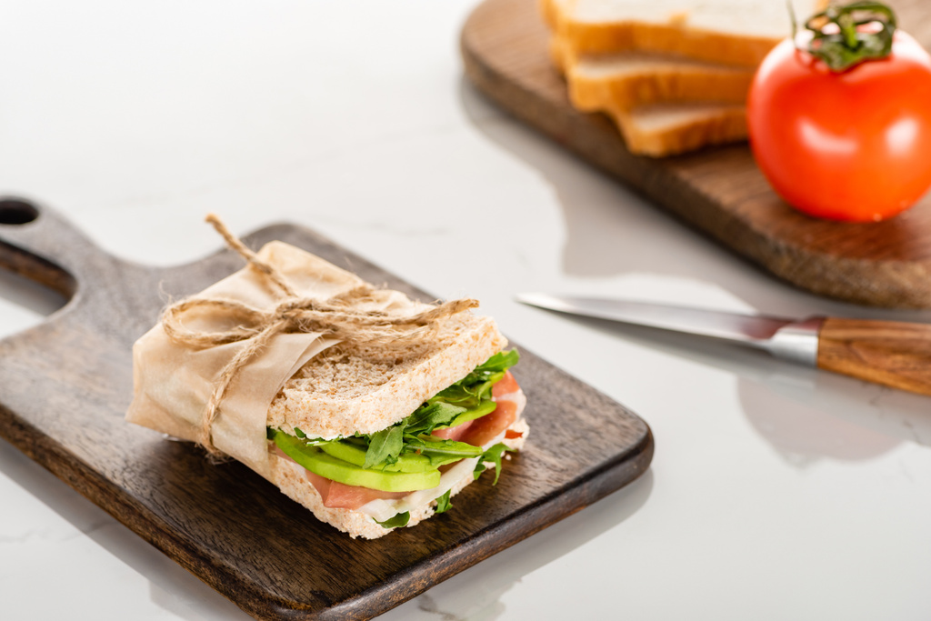 вибірковий фокус свіжого зеленого бутерброда з авокадо і прошутто на дерев'яній обробній дошці на білій мармуровій поверхні
 - Фото, зображення