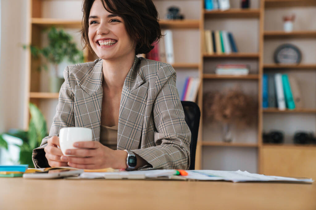 Hermosa joven empresaria sonriente sentada en el escritorio de la oficina, sosteniendo una taza de café
 - Foto, imagen