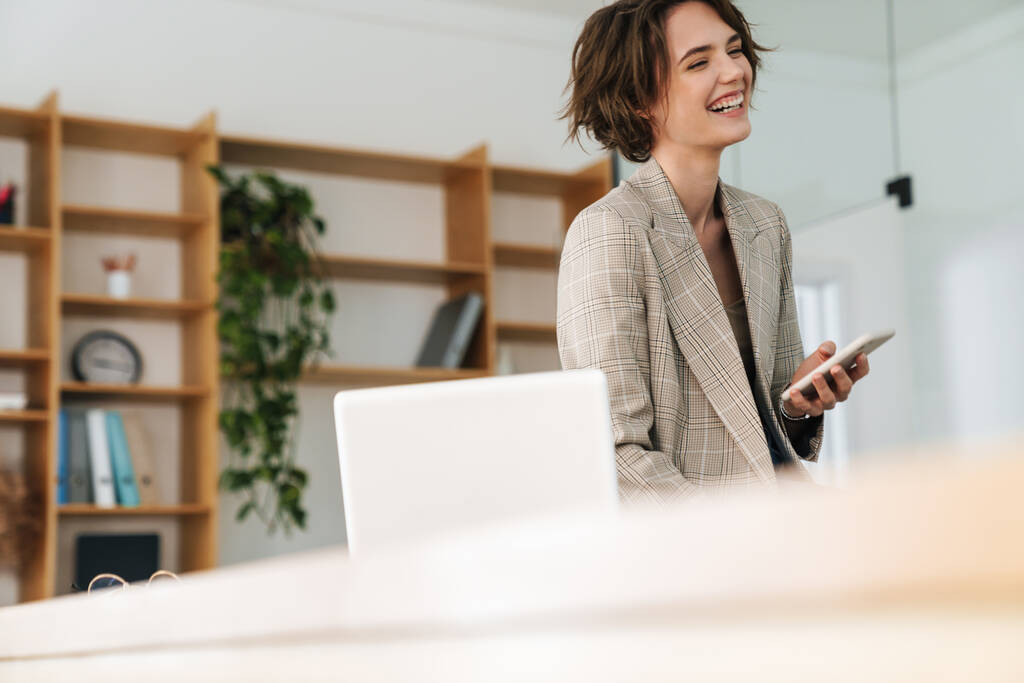 Aantrekkelijke glimlachende jonge zakenvrouw die een jas draagt met een mobiele telefoon terwijl ze op een bureau zit en een laptop op kantoor gebruikt - Foto, afbeelding