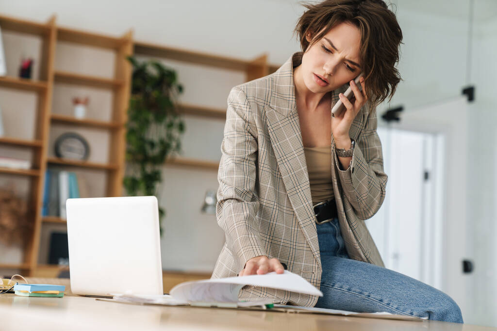 Приваблива впевнена молода бізнес-леді в куртці, що розмовляє на мобільному телефоні, сидячи на столі і використовуючи ноутбук в офісі
 - Фото, зображення