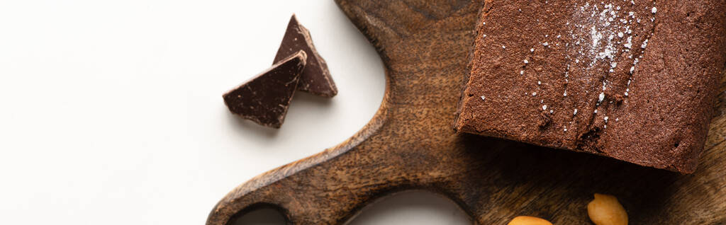 Draufsicht auf köstliches Brownie-Stück auf Holzschneidebrett mit Schokolade auf weißem Hintergrund, Panoramaaufnahme - Foto, Bild