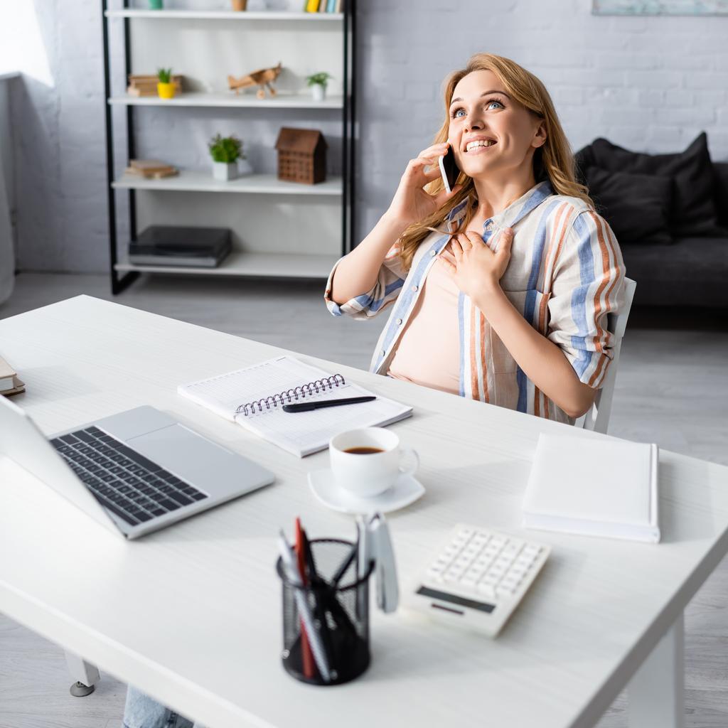 Szelektív fókusz boldog nő beszél okostelefon közelében laptop, írószer és kávé az asztalon  - Fotó, kép