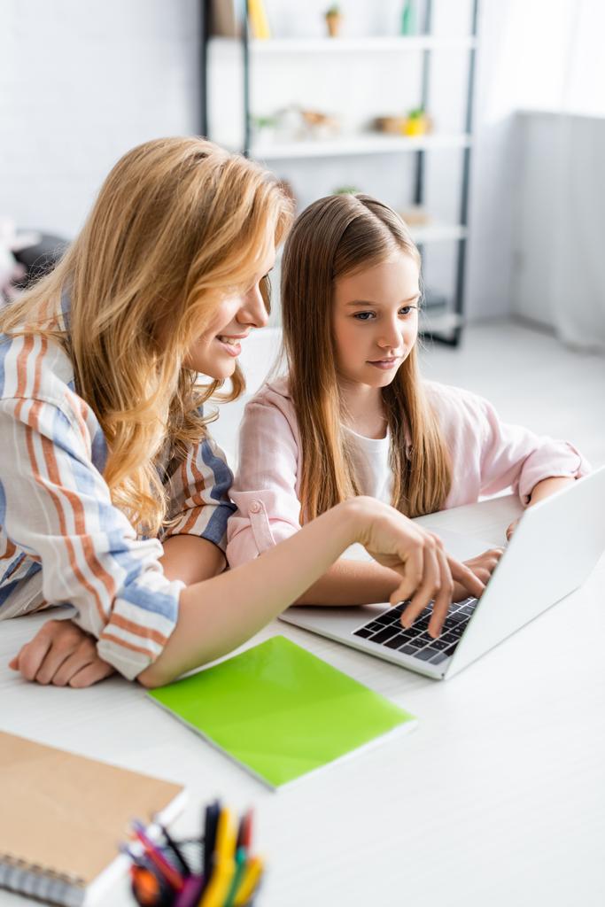 Selectieve focus van lachende moeder met behulp van laptop in de buurt dochter tijdens webinar thuis  - Foto, afbeelding
