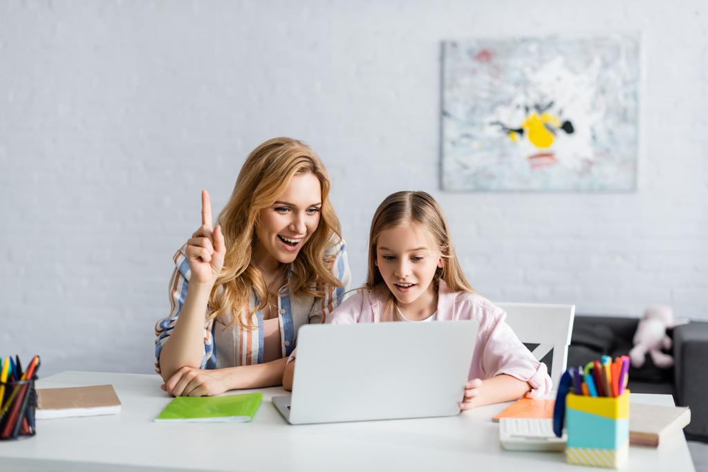 Enfoque selectivo de la mujer sonriente que tiene idea mientras usa el ordenador portátil cerca del niño y artículos de papelería en la mesa
  - Foto, Imagen