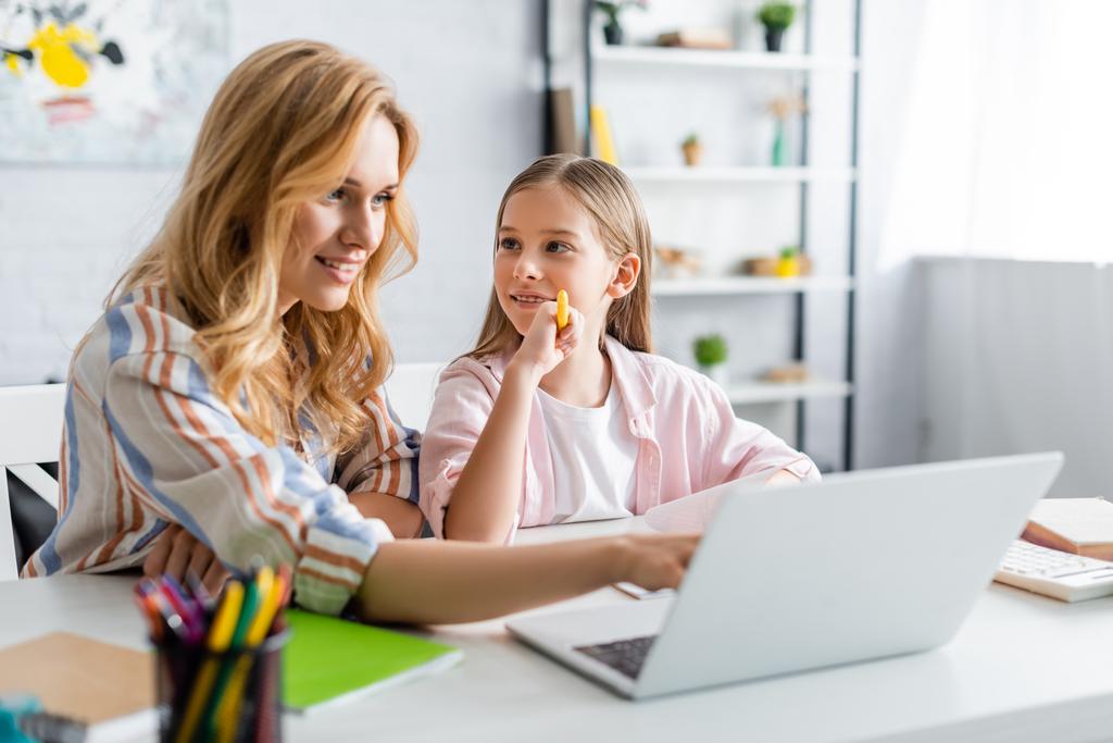 Enfoque selectivo de la mujer sonriente usando el ordenador portátil cerca del niño sosteniendo la pluma durante la educación en línea
  - Foto, Imagen