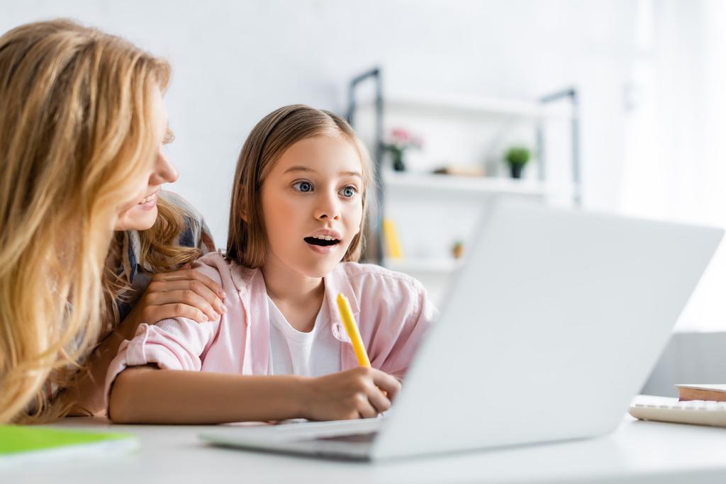 Valikoiva painopiste hymyilevä nainen syleilee tytär kirjallisesti kannettavan aikana online-koulutuksen kotona  - Valokuva, kuva