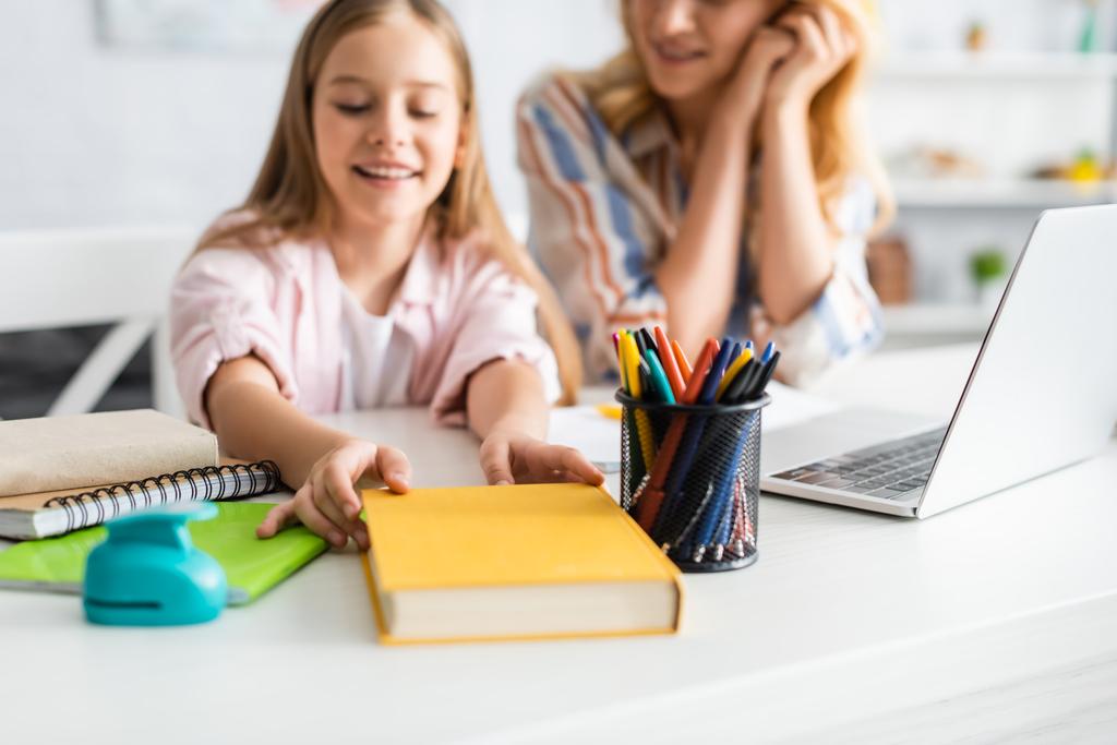 Вибірковий фокус усміхненої дитини, що бере книгу біля матері та ноутбука на столі
  - Фото, зображення
