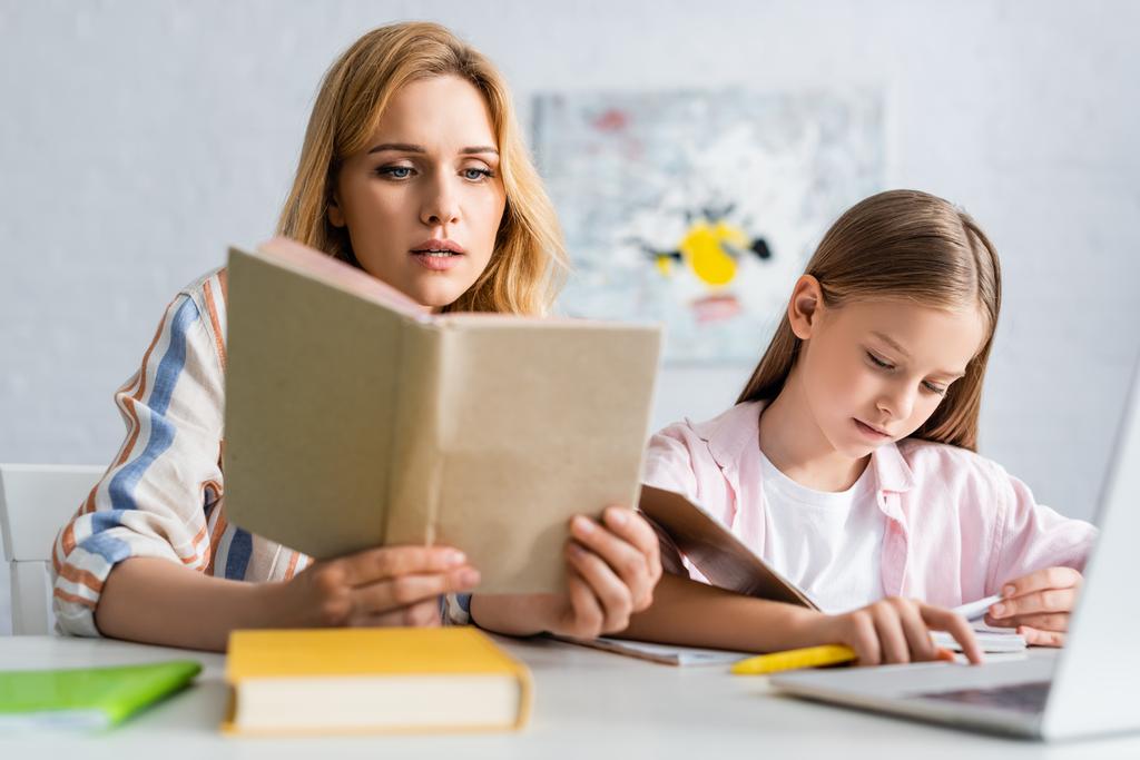 Enfoque selectivo de la mujer leyendo libro mientras ayuda a su hija durante la educación en línea en el hogar
  - Foto, Imagen