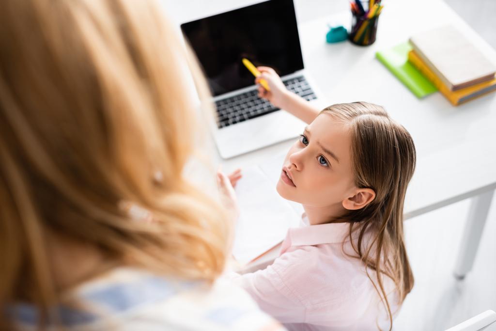 Vue aérienne de l'enfant regardant sa mère tout en pointant vers un ordinateur portable pendant l'éducation en ligne  - Photo, image
