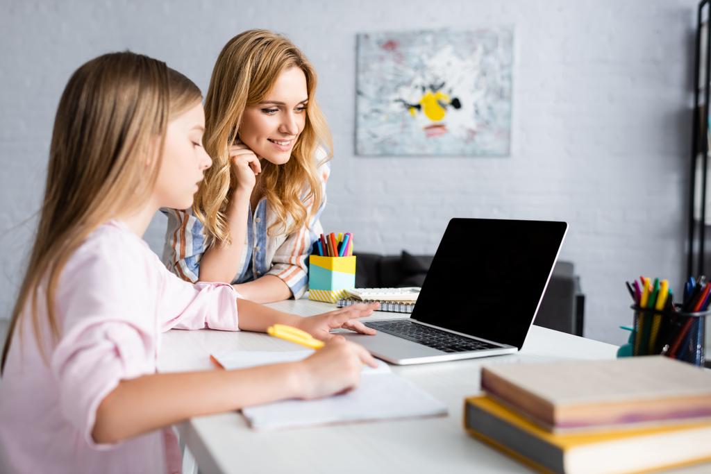 A mosolygós nő szelektív fókusza, aki a gyerek mellett áll, és otthon, elektronikus tanulás közben laptopot használ  - Fotó, kép