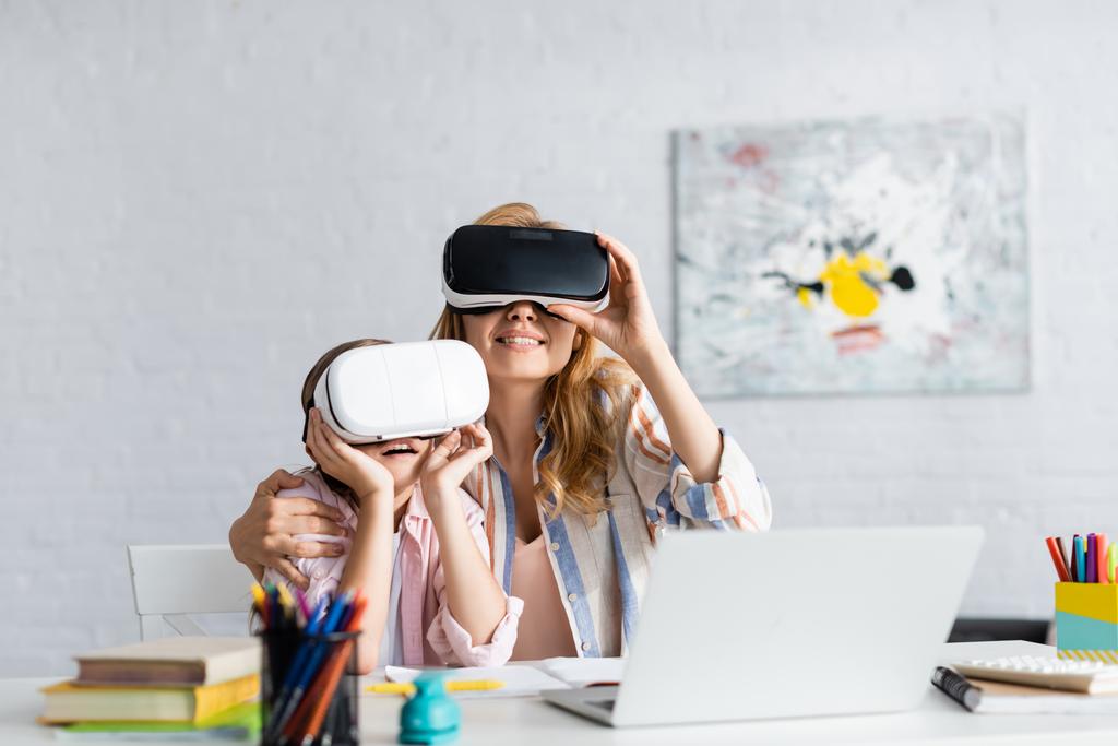 Szelektív fókusz mosolygós nő átölelő lánya használata közben virtuális valóság headset közelében laptop és könyvek  - Fotó, kép