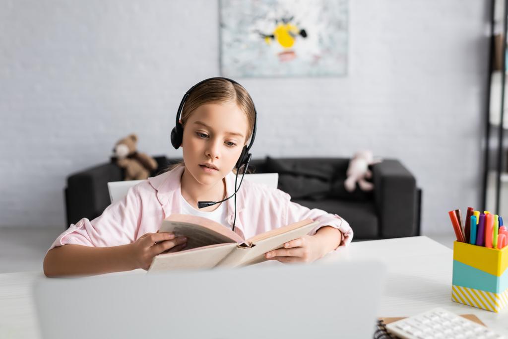 Concentration sélective de l'enfant dans le livre de lecture du casque près de l'ordinateur portable sur la table  - Photo, image