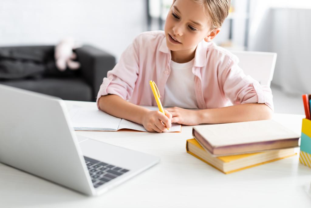 Foco seletivo de criança bonito escrevendo em notebook perto de livros e laptop durante a educação eletrônica
  - Foto, Imagem