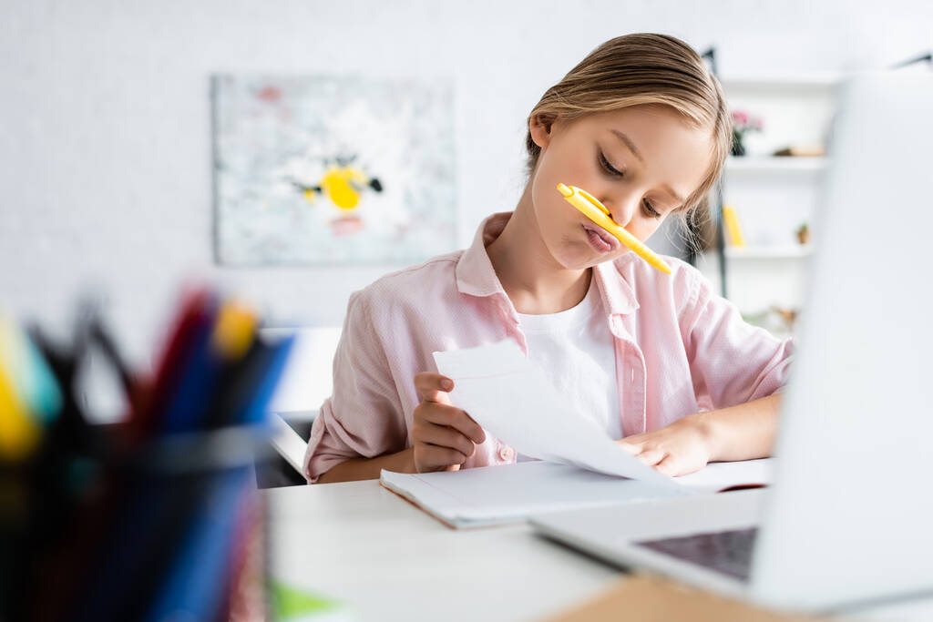 Селективный фокус ребенка с ручкой возле рта, глядя на ноутбук рядом с ноутбуком
  - Фото, изображение
