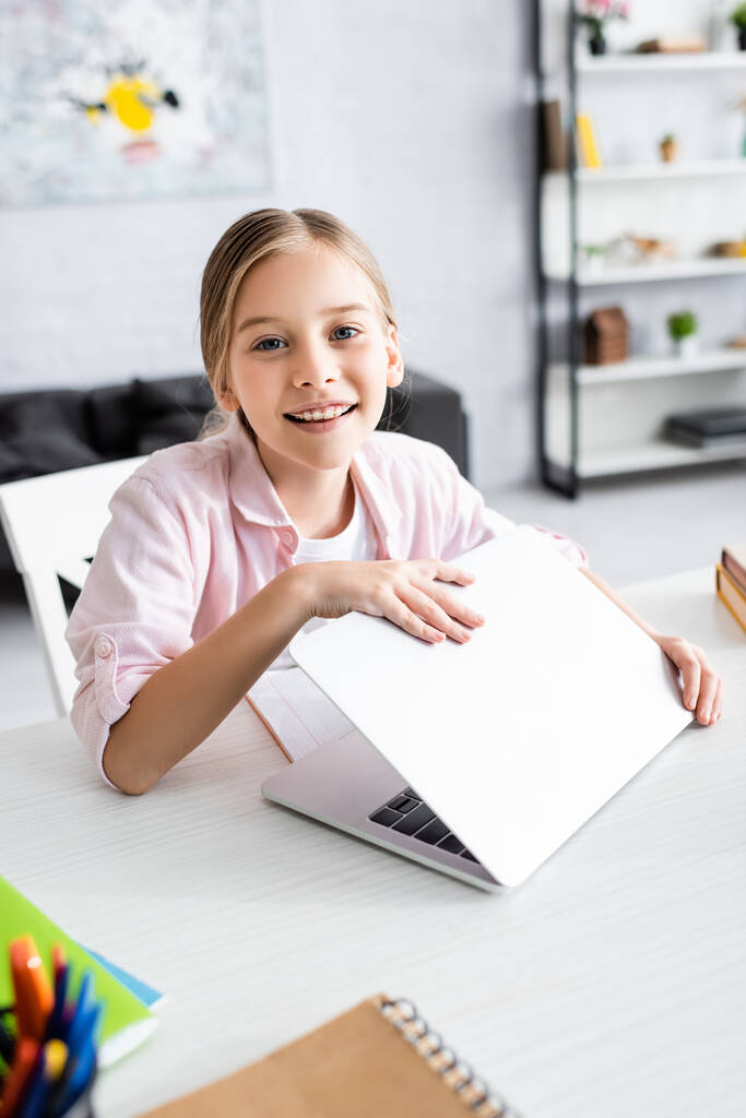 Foco seletivo de criança sorrindo olhando para a câmera enquanto segurando laptop perto de artigos de papelaria na mesa
  - Foto, Imagem