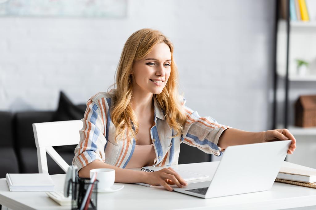 Selective focus of smiling woman using laptop at desk - Photo, Image