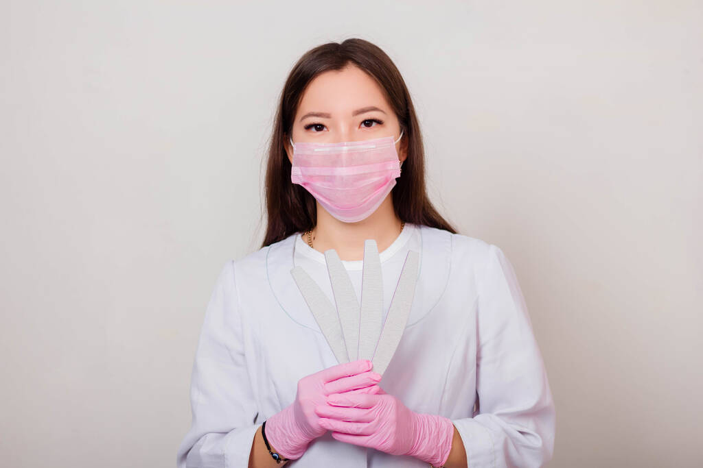 Portrait of a woman master of nails, in a white coat and pink mask on her face with nail files in her hands. Photo on a white background. - Photo, Image