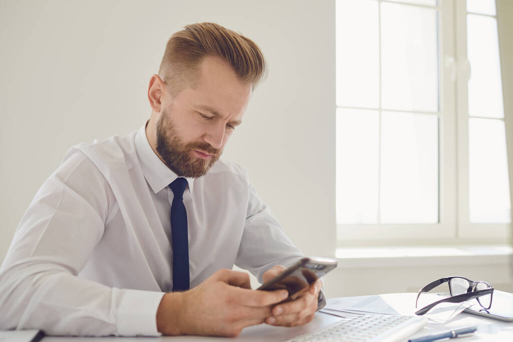 Серьезный занятой бизнесмен читает сообщение по телефону за столом с компьютером в офисе
. - Фото, изображение