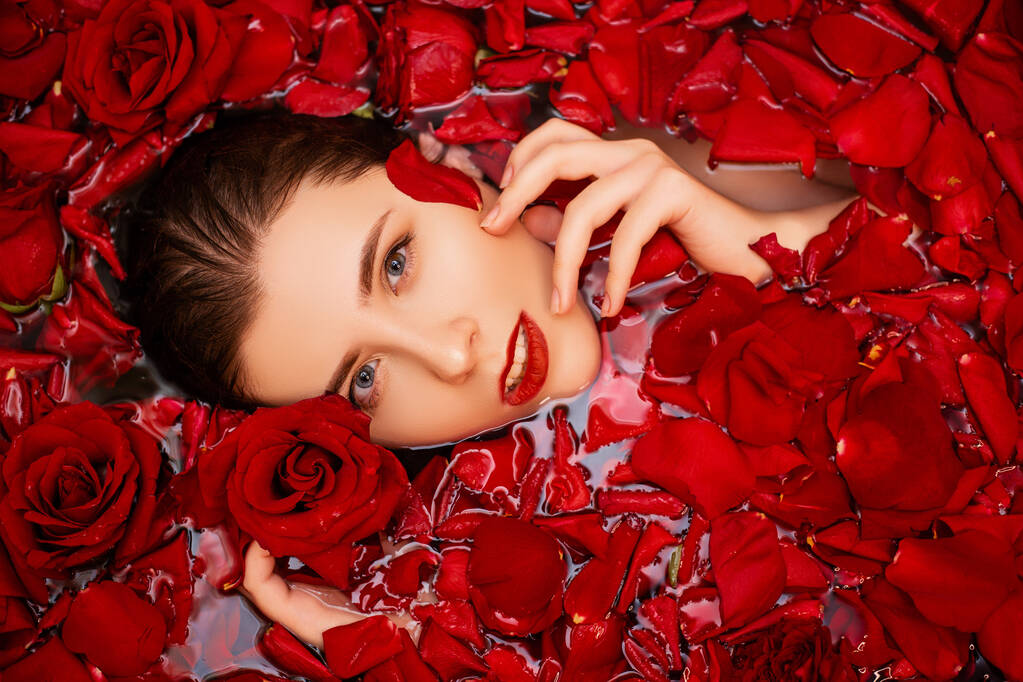 Vista superior, rosto close-up de uma jovem mulher em uma banheira cheia de pétalas de rosa vermelha. Retrato de beleza de uma menina atraente. Tratamento de spa para rejuvenescimento da pele
. - Foto, Imagem