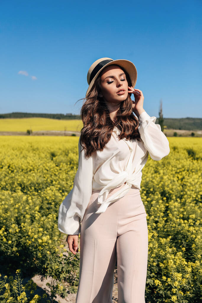 photo extérieure de mode de belle femme aux cheveux foncés dans des vêtements élégants et chapeau de paille posant dans le champ de colza en fleurs - Photo, image