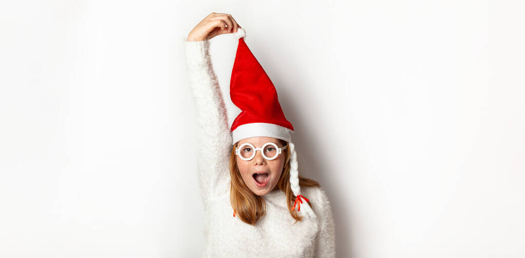 新年の感情と混乱サンタクリスマスの帽子の女の子は白い背景に隔離された彼の手で帽子を持っています. - 写真・画像