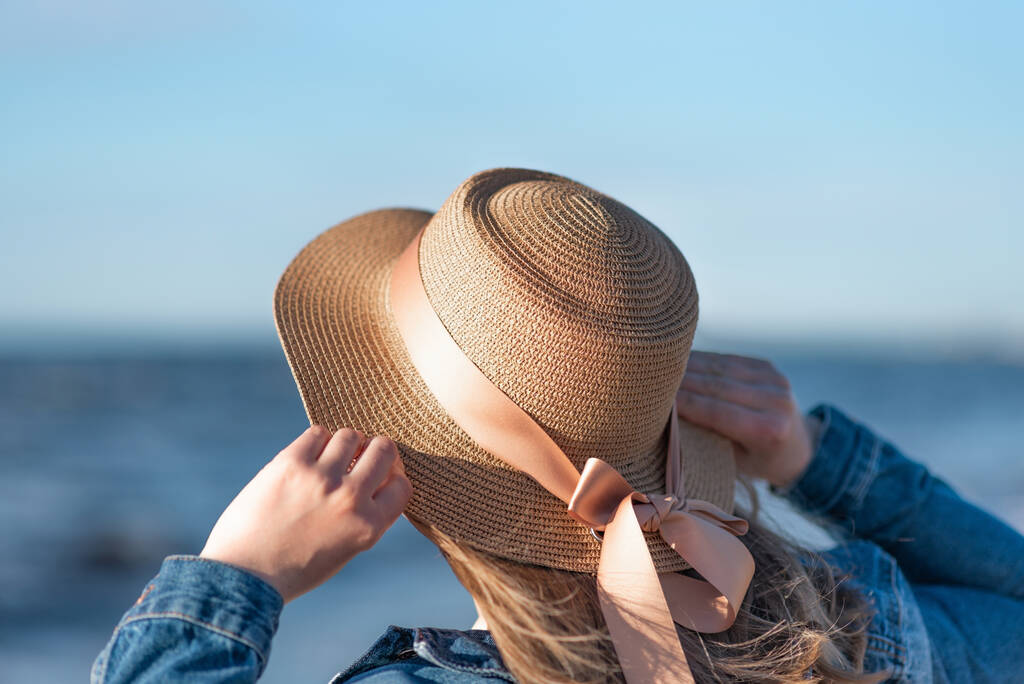 donna in un cappello di paglia e giacca di jeans su uno sfondo del mare. Vista posteriore da vicino - Foto, immagini