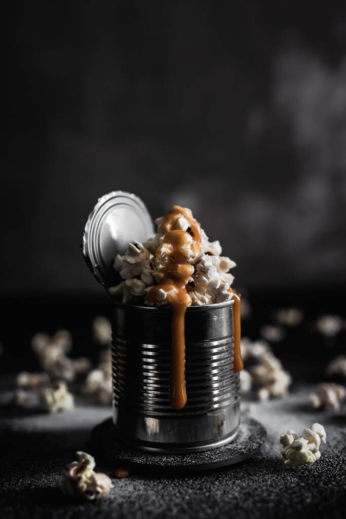 Une boîte en acier de maïs soufflé au caramel salé sur fond sombre, photo lunatique - Photo, image
