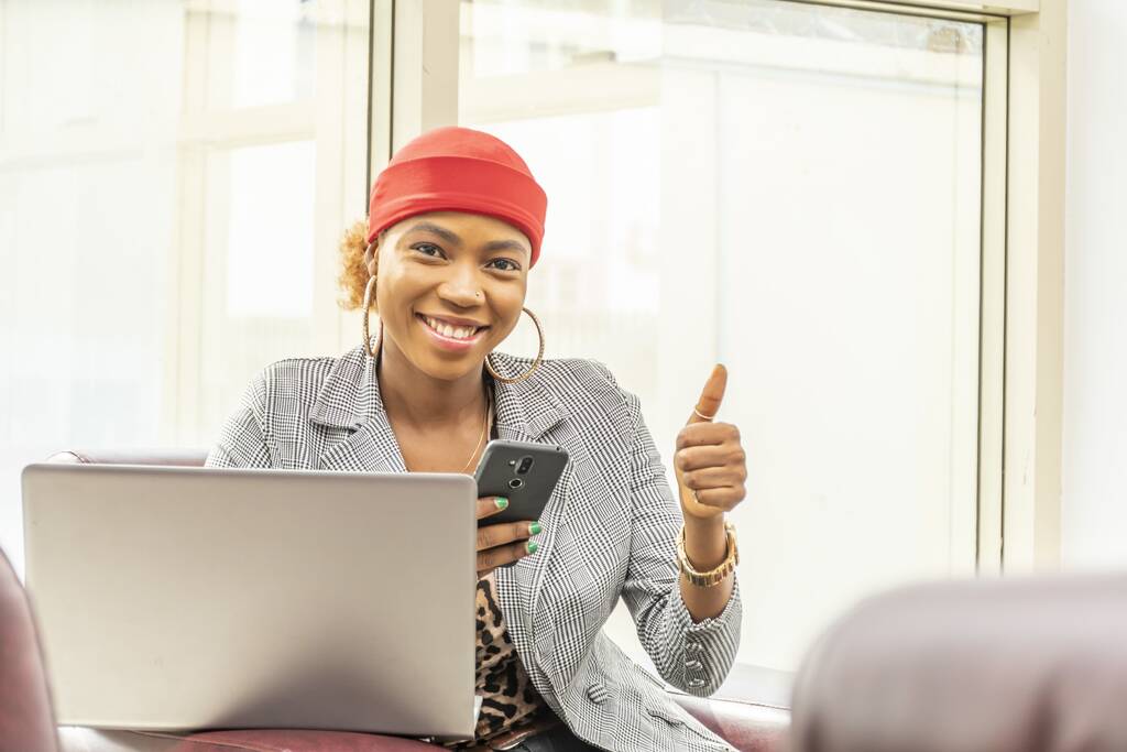 Eine Afroamerikanerin sitzt auf einem Sessel und macht die Daumen-hoch-Geste, während sie einen Laptop und ein Smartphone benutzt - Foto, Bild