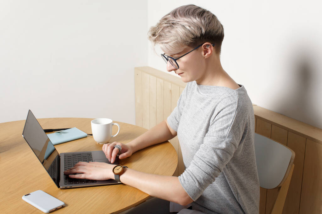 Młoda hipsterka skupiona millenium kobieta z krótką blond fryzurą pracuje z laptopem we wnętrzu kawiarni, widok z bliska, patrząc na ekran i wpisując - Zdjęcie, obraz