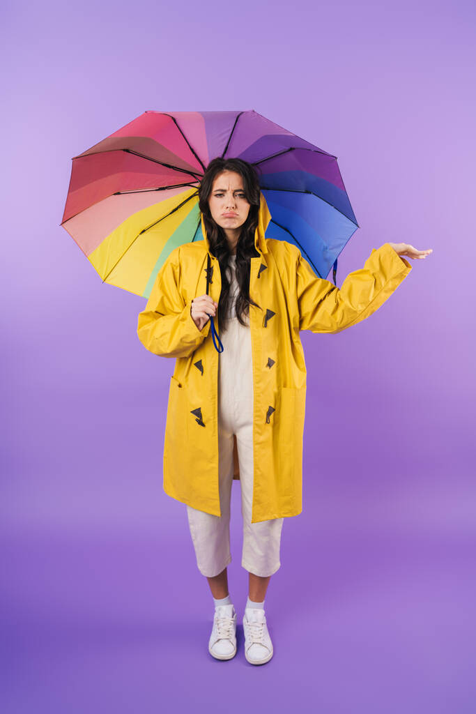 Photo of cute sad brunette woman in yellow raincoat posing isolated over purple wall background holding umbrella. - Photo, Image