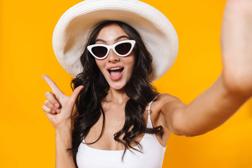 Image of excited woman in swimsuit and hat pointing finger aside while taking selfie photo isolated over yellow wall - Photo, Image